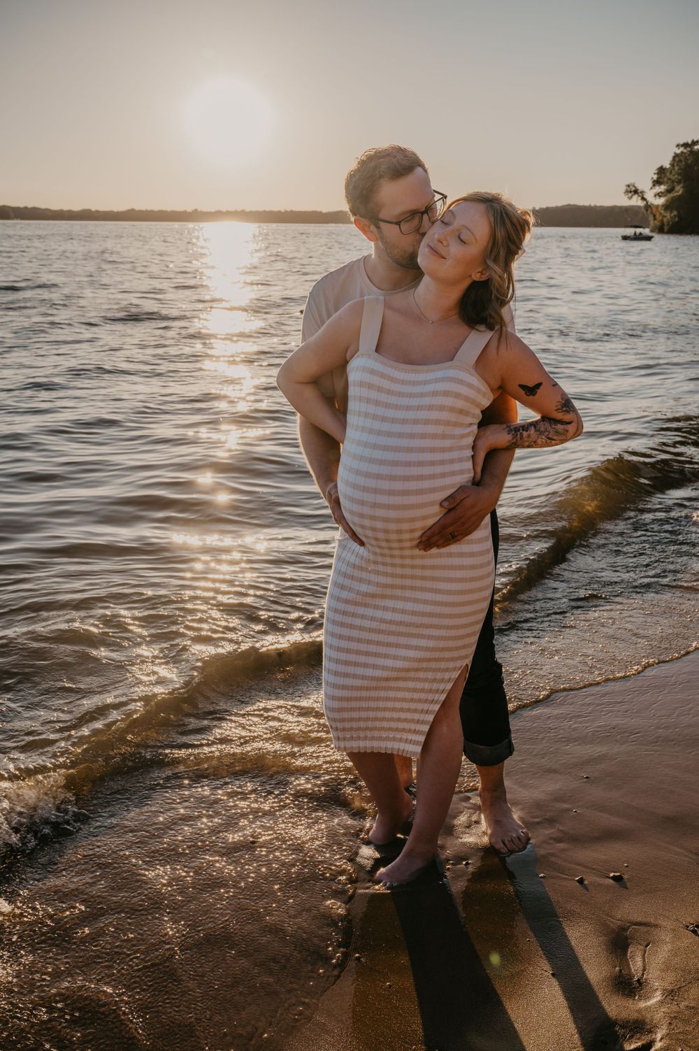 Couple embracing on a sandy beach at sunset, man holds pregnant woman's belly in warm golden light.