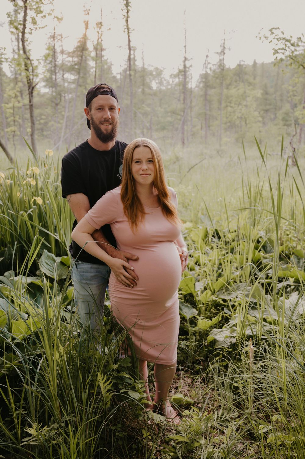Expecting couple stands in lush green meadow, woman in pink maternity dress cradling baby bump at golden hour.