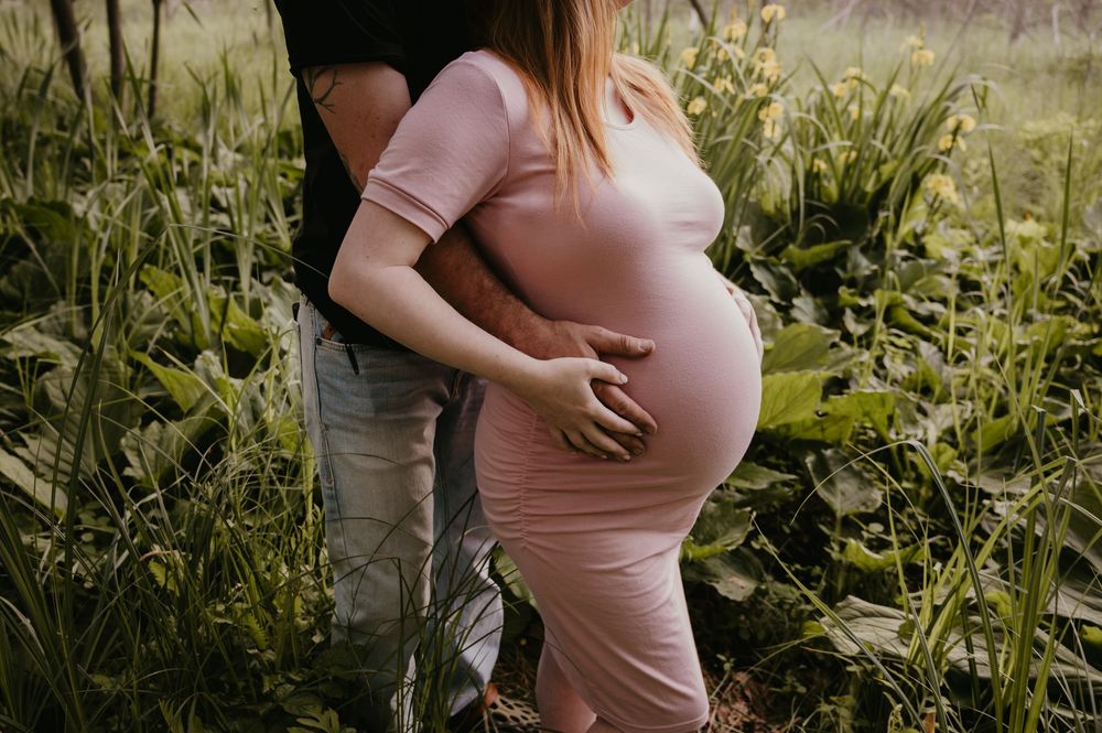 Close-up of pregnant woman in pink dress with partner embracing her from behind surrounded by green foliage.