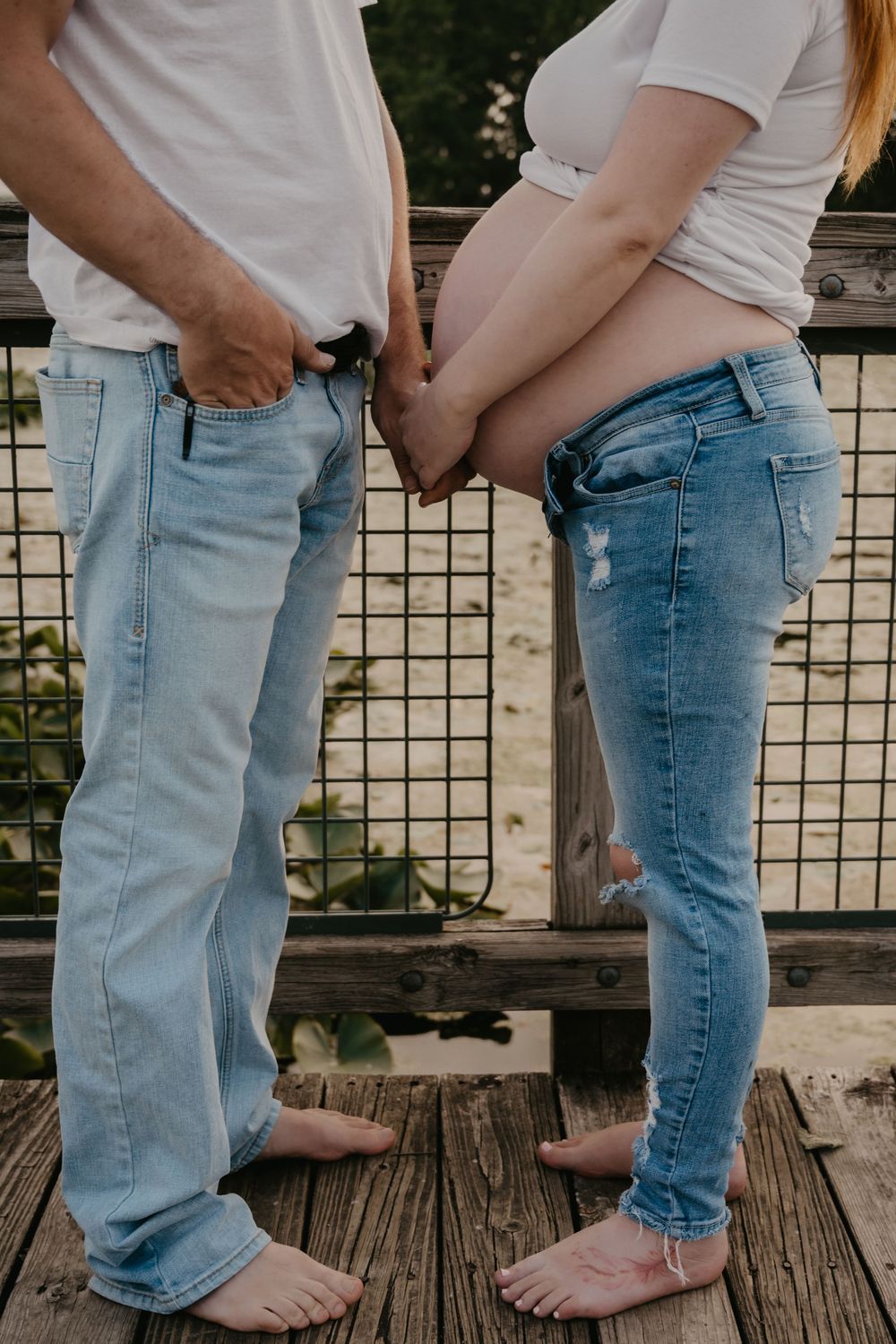 Barefoot expecting couple stands on wooden bridge holding hands, woman in ripped jeans showing bare baby bump.