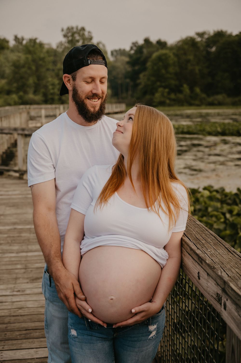 Bearded man in white tee smiles at pregnant redhead partner on wooden bridge at sunset, both cradling baby bump.