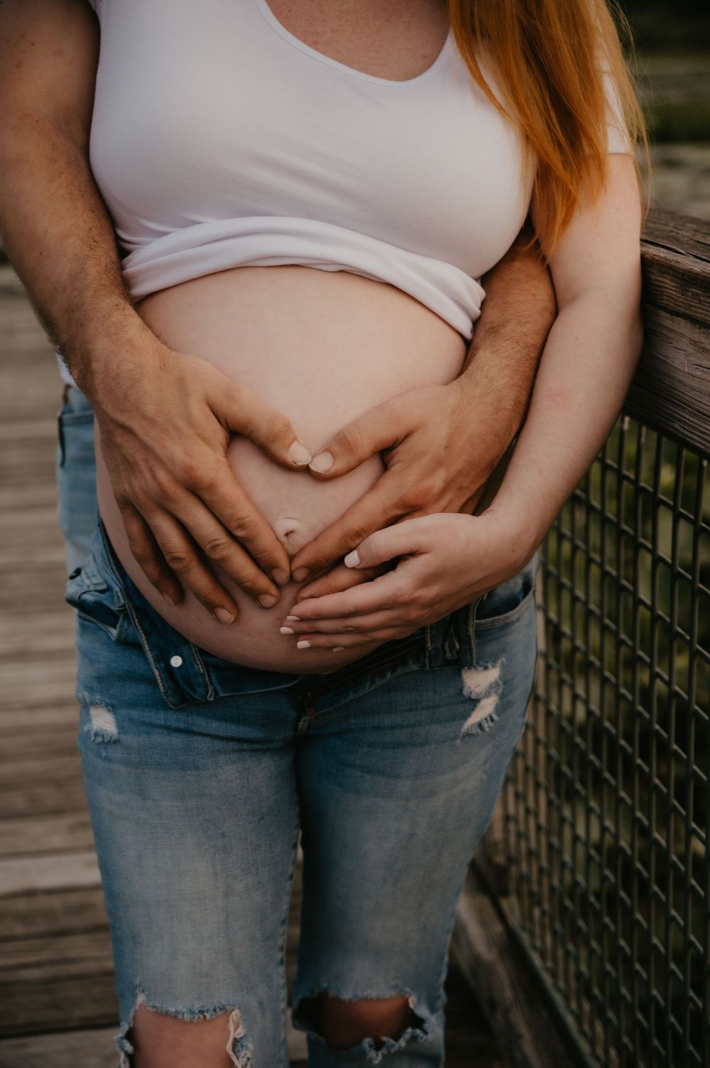 Couple forms heart shape with hands on pregnant belly, woman in white tank and ripped jeans on wooden bridge.