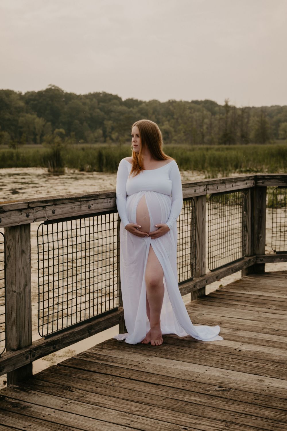 Pregnant woman in flowing white maternity gown standing on wooden dock by serene lake surrounded by lush greenery.