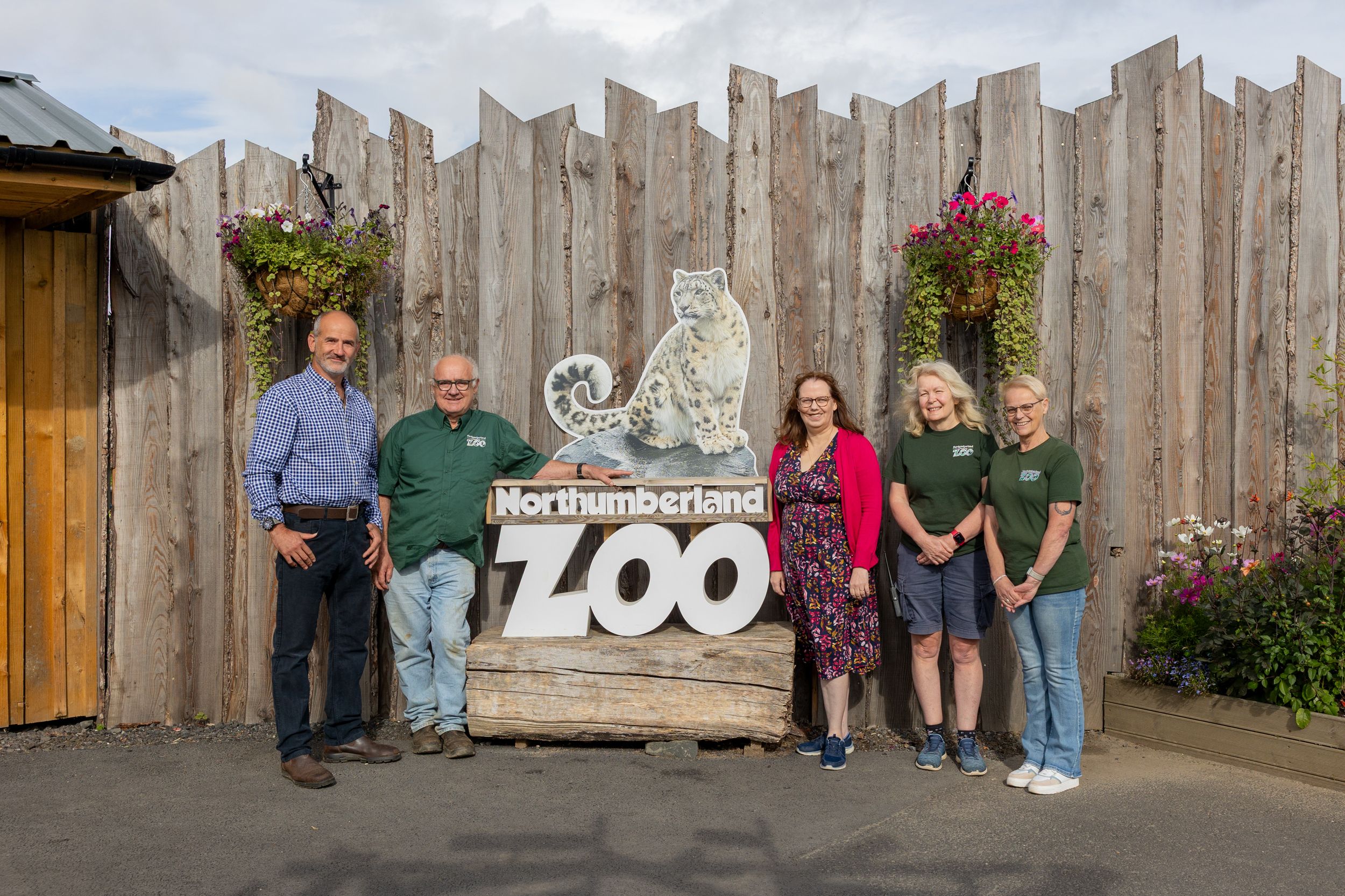 Five people posing in front of the Northumberland Zoo entrance sign with hanging flower baskets and wooden fence backdrop.
