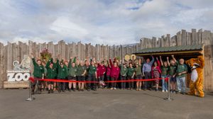 Large group of people in green uniforms celebrate a ribbon-cutting ceremony outside a zoo entrance with a tiger mascot.