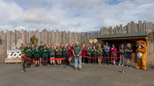 Zoo staff and guests gather for a ribbon-cutting ceremony outside a wooden zoo entrance with a mascot present.