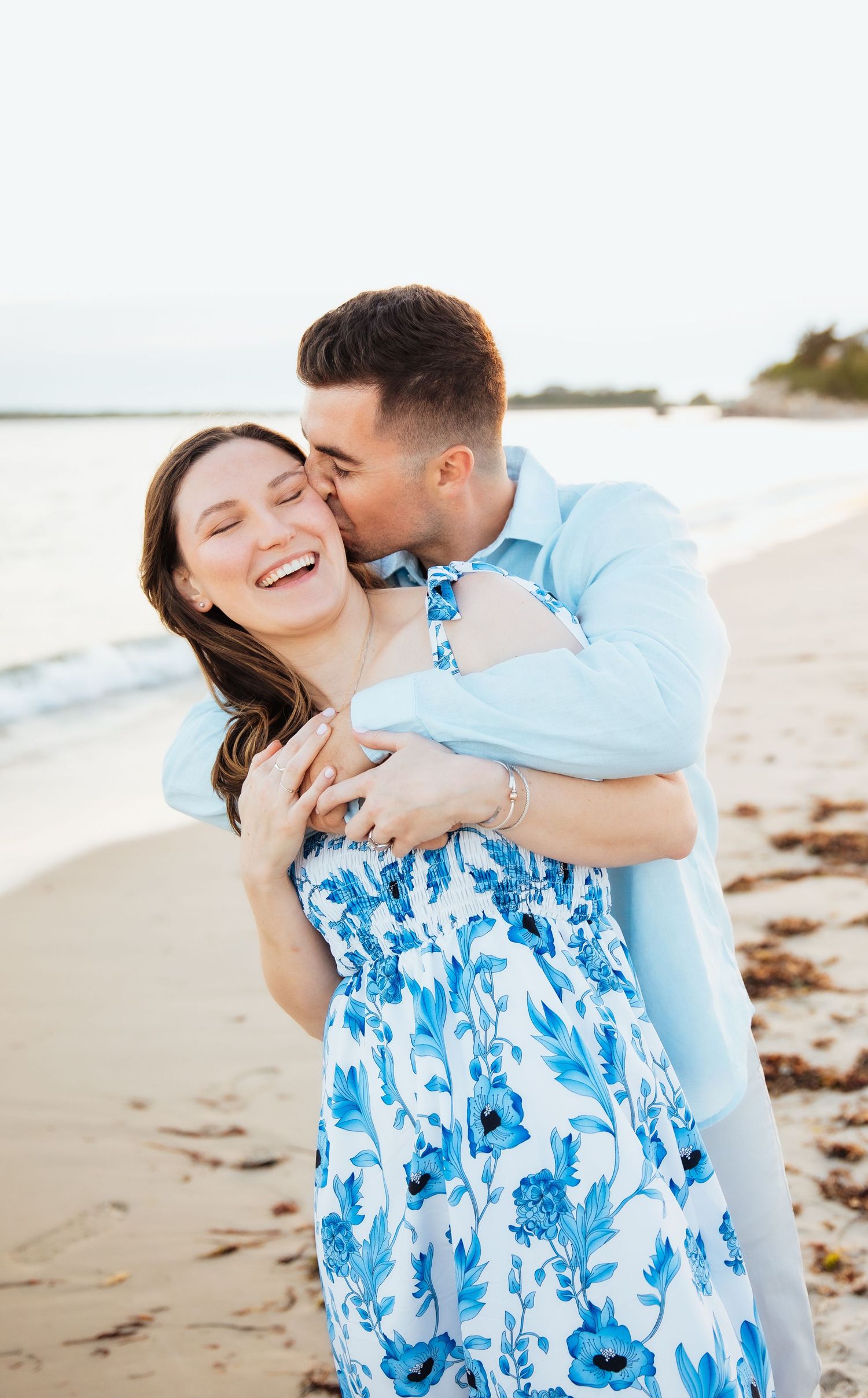 Couple sitting on beach with their dog wearing blue bandana at sunset. Engagement photoshoot