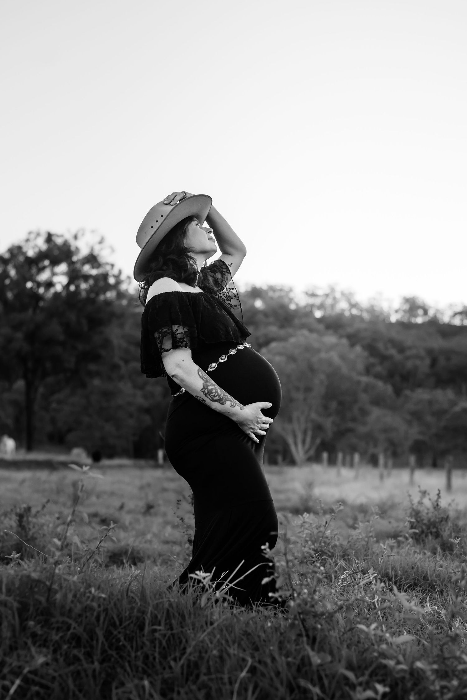 Black and white maternity portrait of woman in hat standing in field.