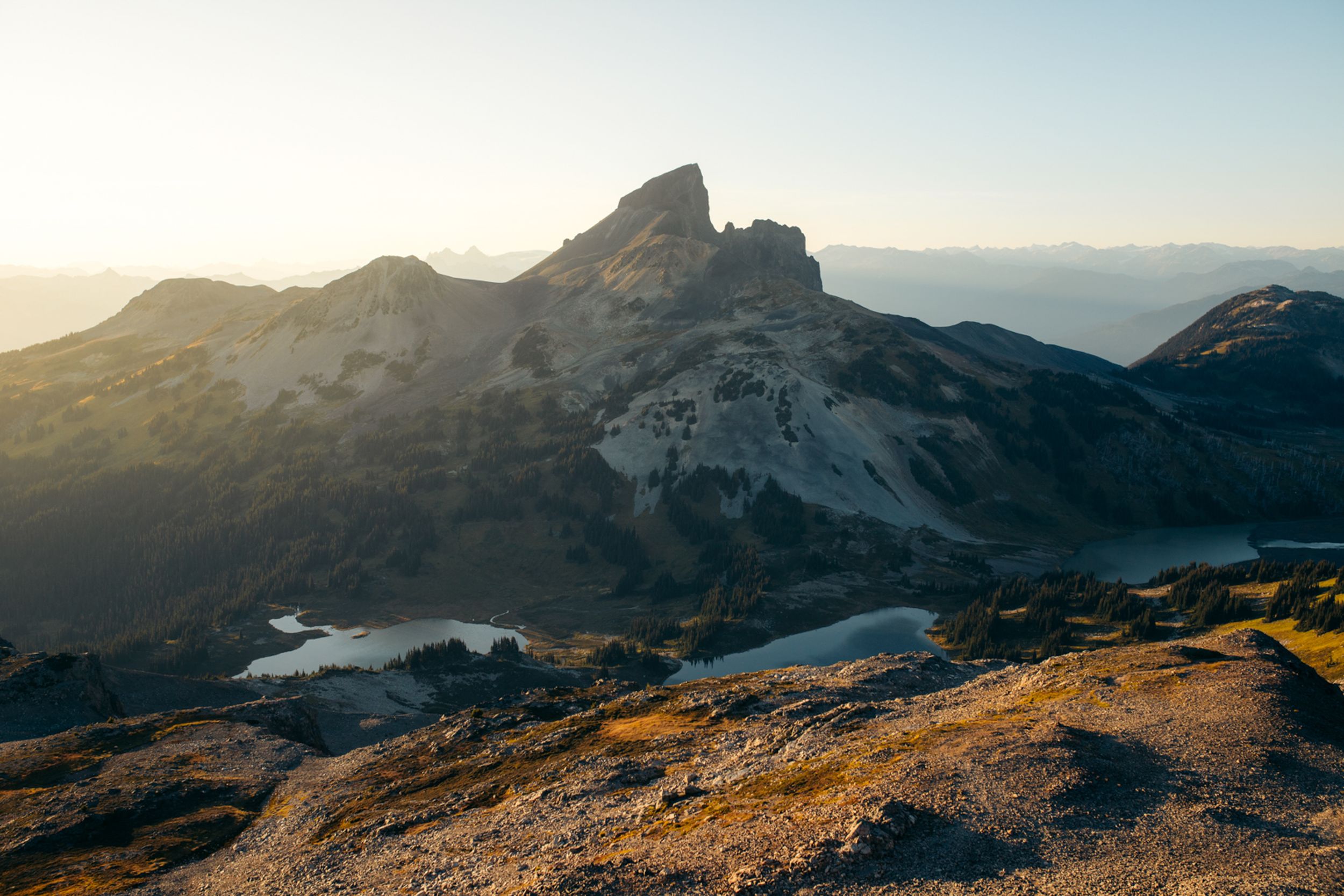 Rugged mountain peaks catch golden sunlight at sunset with rocky terrain and patches of snow in the foreground.