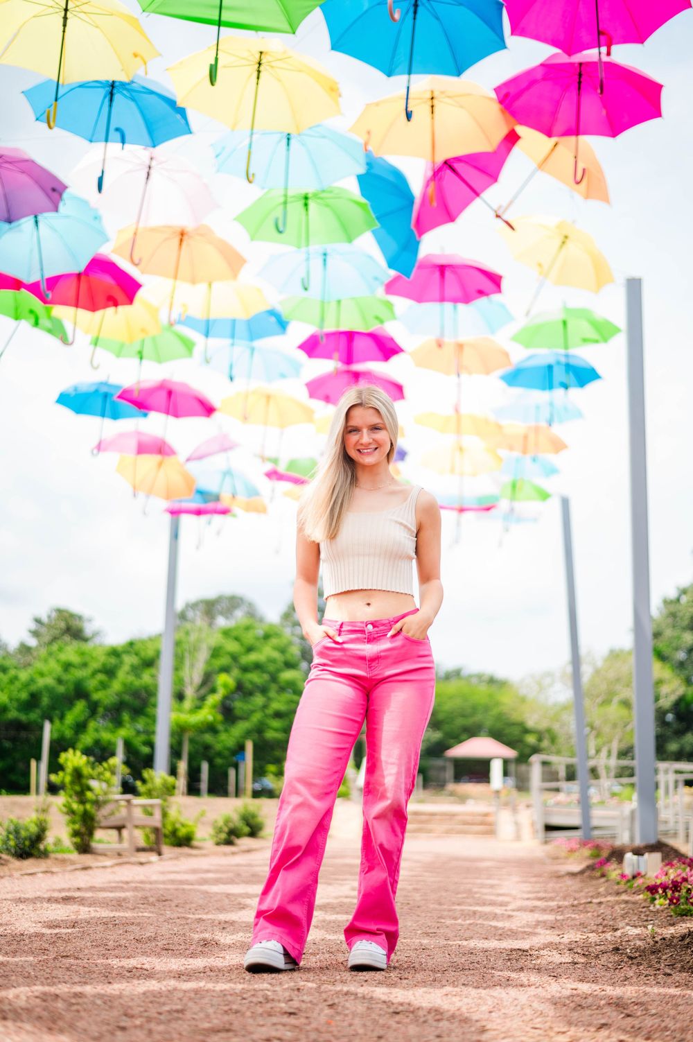 A fashion portrait under colorful suspended umbrellas wearing pink pants and a cream top.
