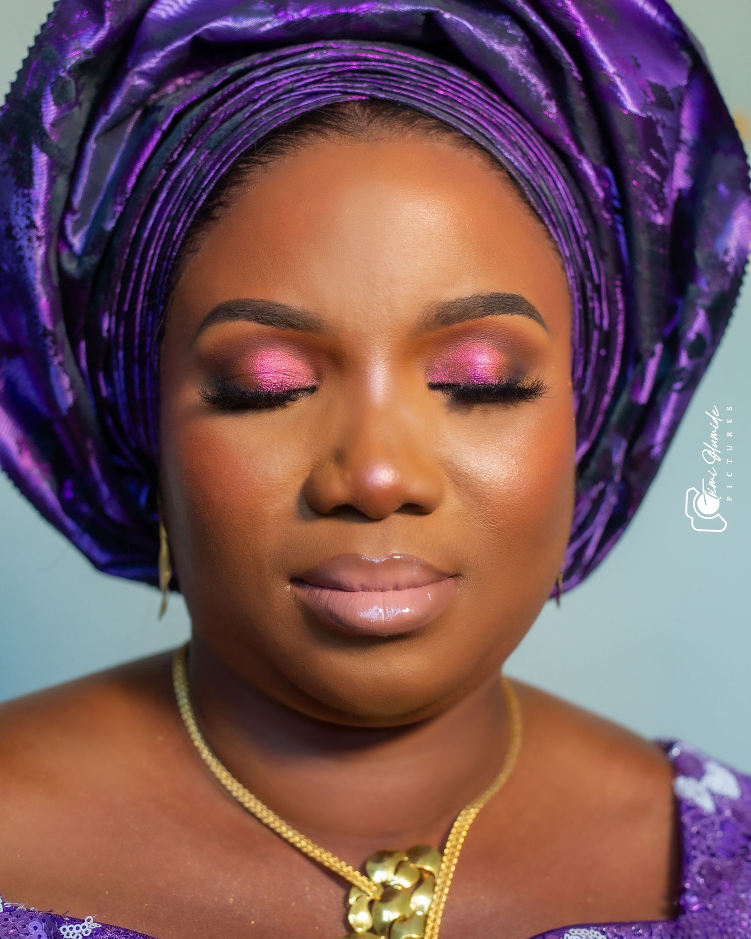 Close-up beauty shot featuring purple gele headwrap and pink eyeshadow with gold necklace.