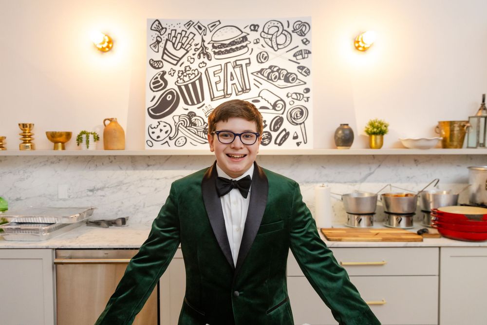 Person in green tuxedo poses in kitchen with food-themed artwork on wall.