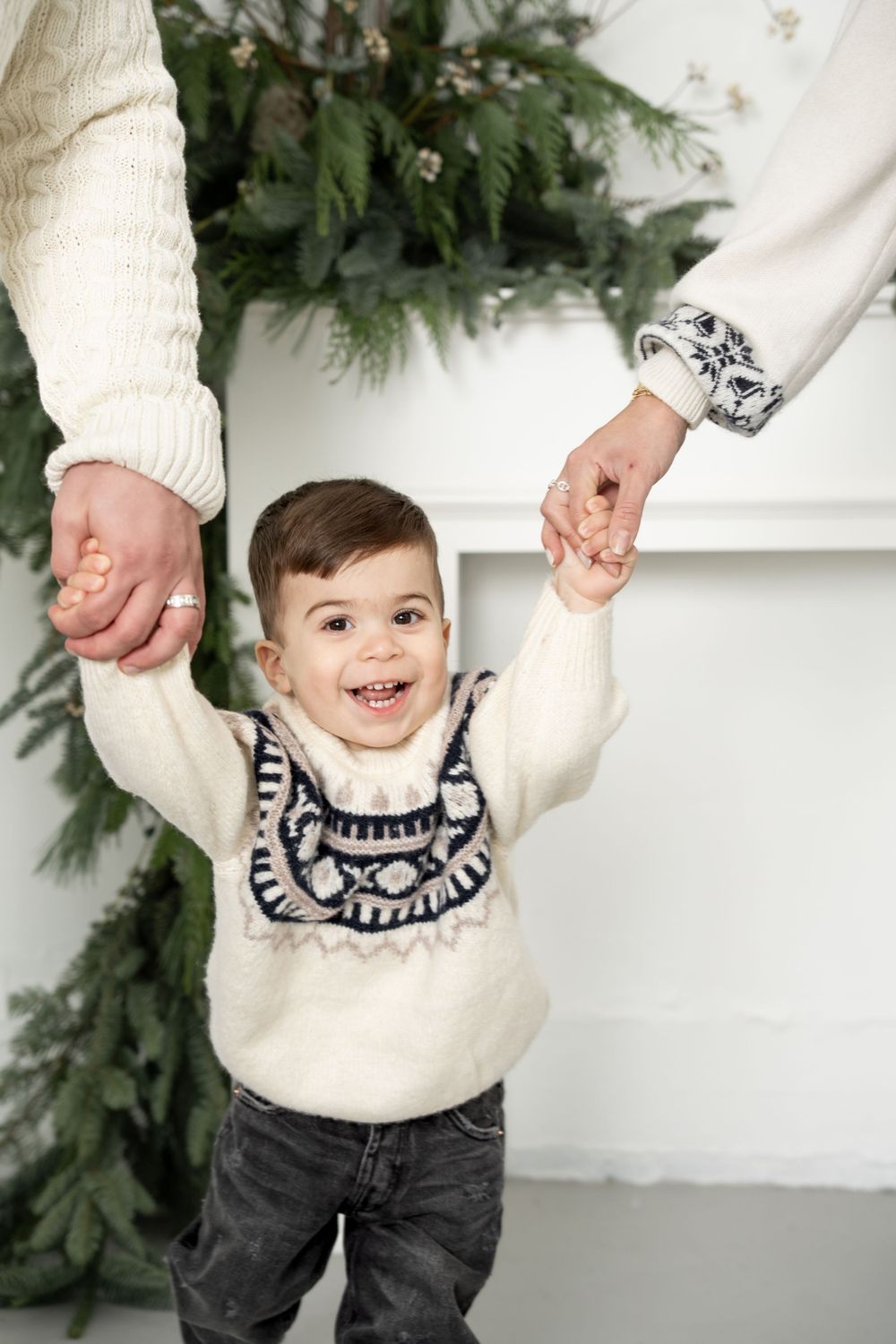 Child wearing cream sweater with navy pattern holds hands with adults against white wall with greenery decor.