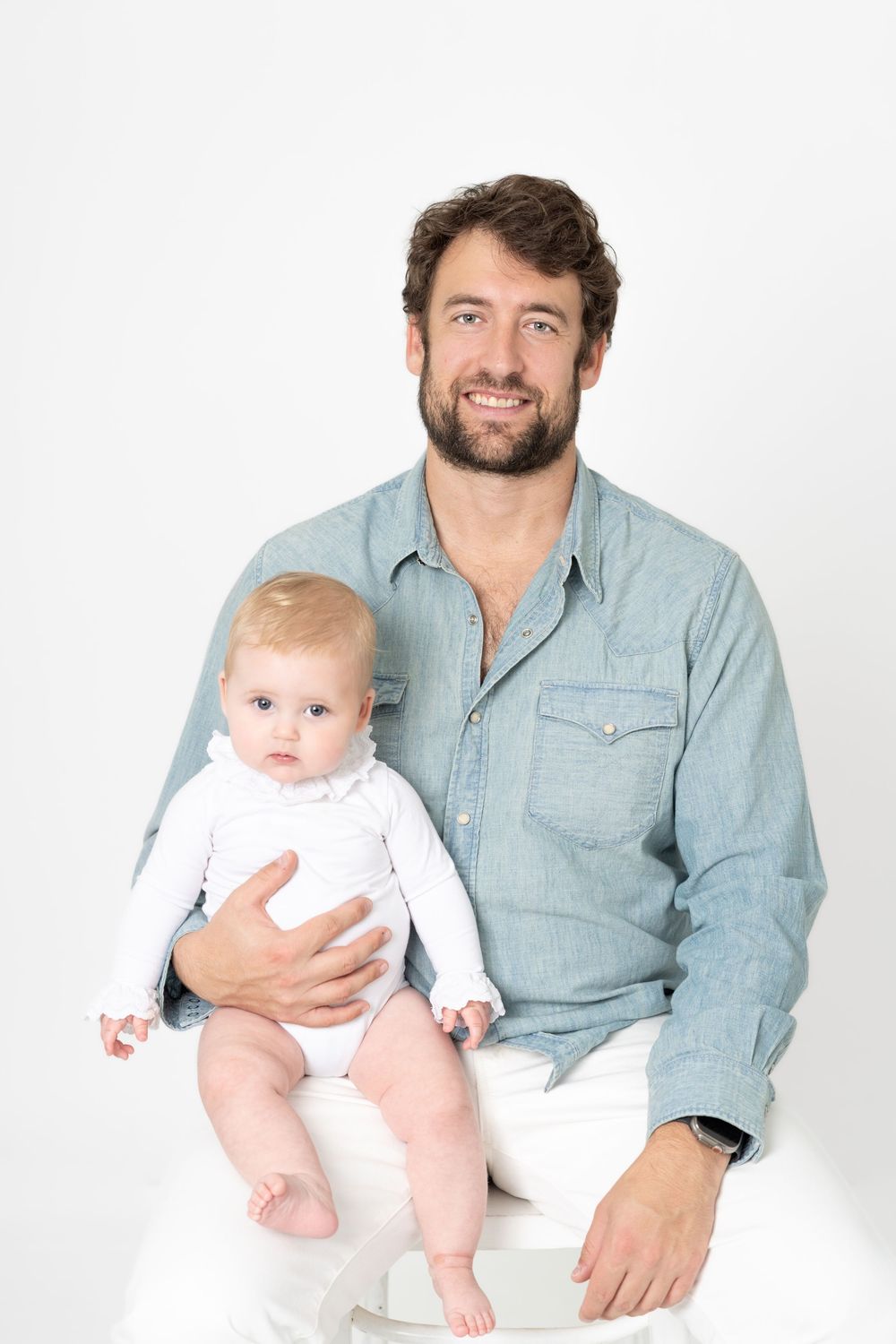 Candid family portraits showing tender moments between parents and baby in casual attire against white backdrop.