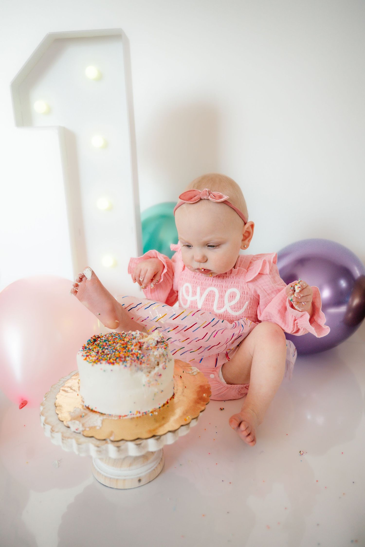 A cute baby in a pink one romper reaches into her first birthday sprinkle smash cake.