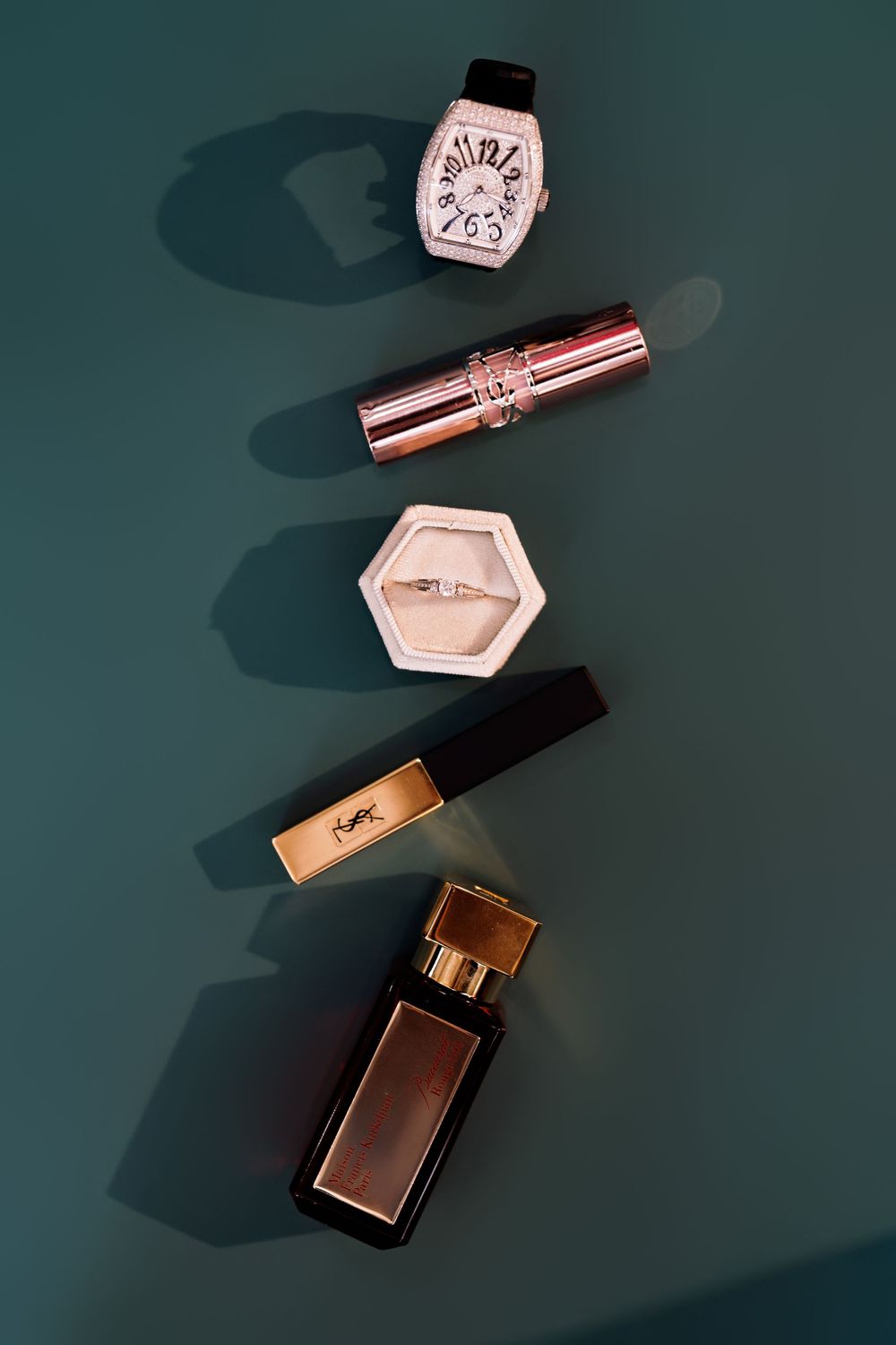 Luxury beauty products including a watch, lipstick, cosmetics and perfume arranged on a dark teal background.