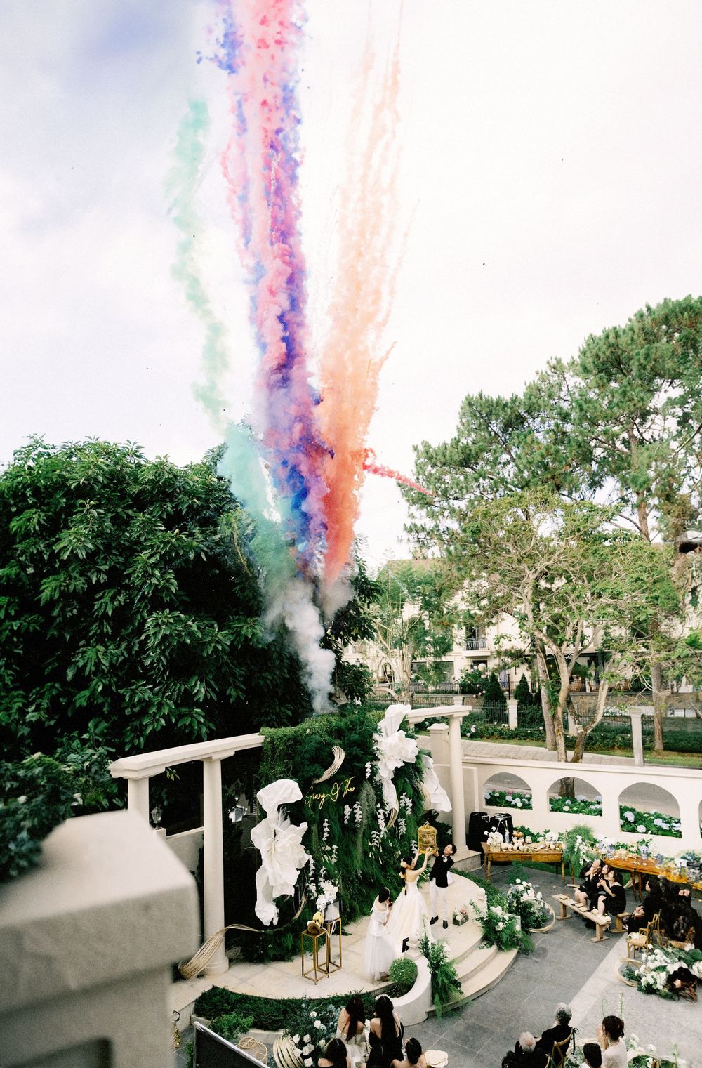 Colorful smoke trails burst into the sky above a decorated outdoor venue with trees and greenery in the background.