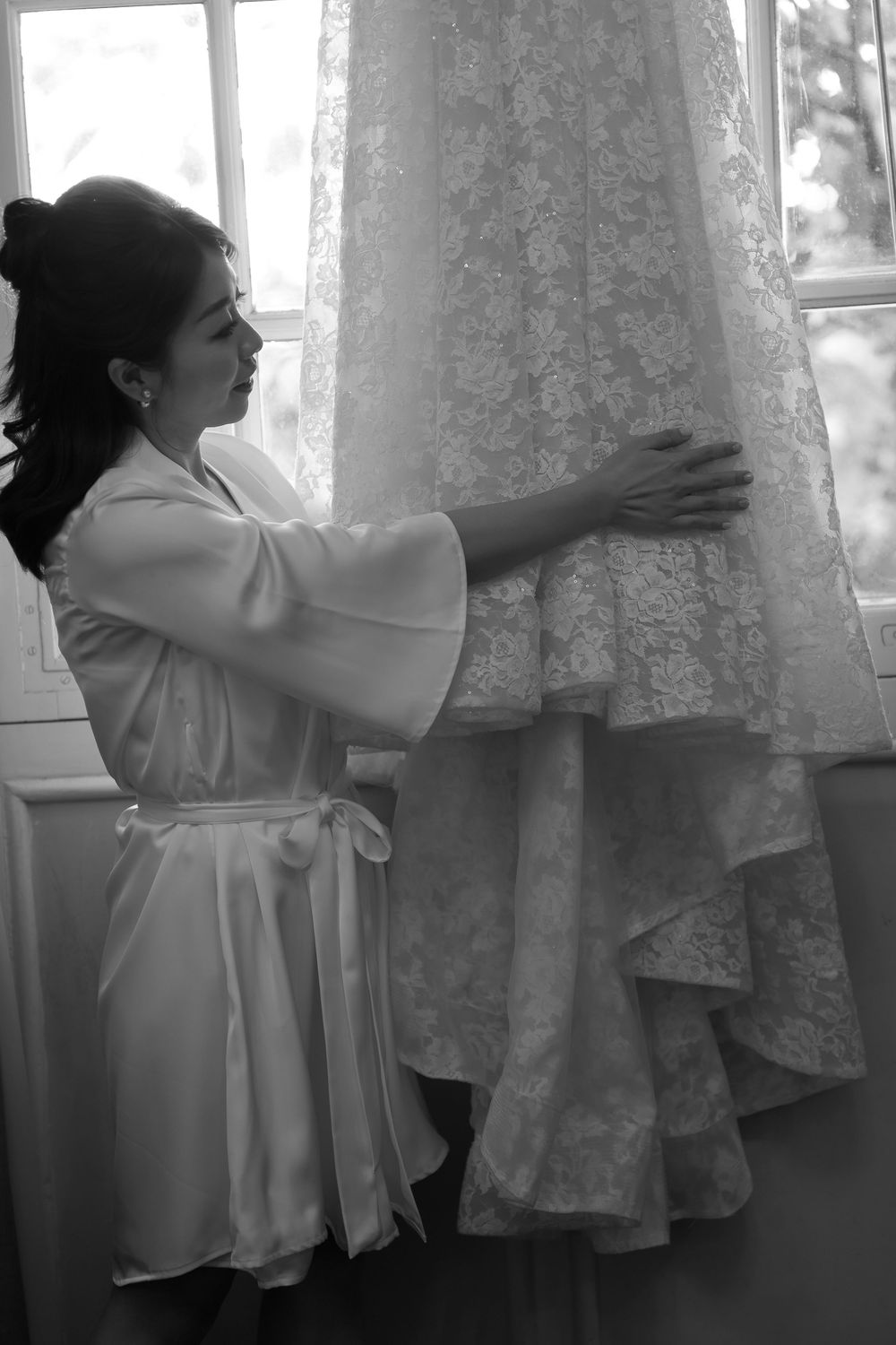 A person in a white robe reaches up to touch an elegant lace wedding dress hanging by a window in black and white.