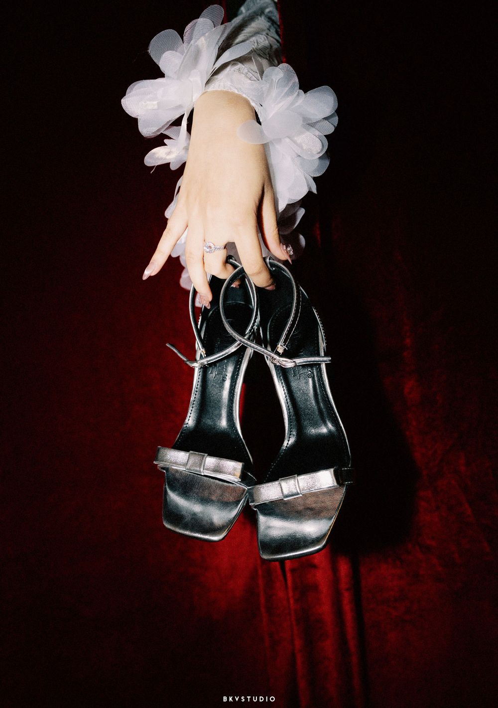 A pale hand holding black platform sandals adorned with white flowers against a dark red background.