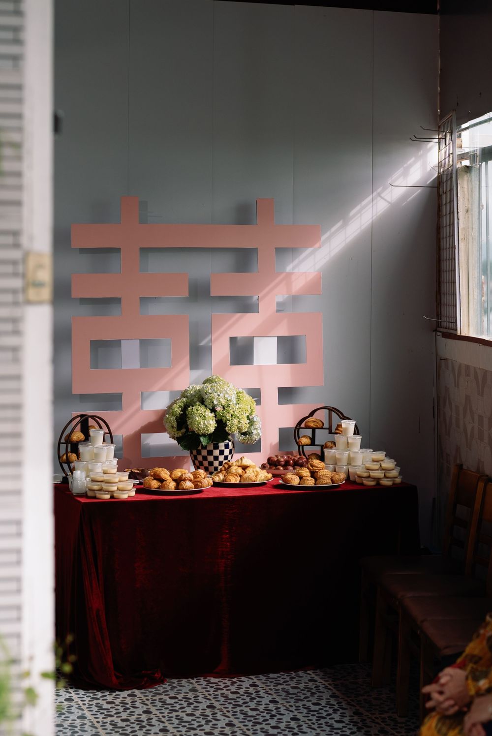 Traditional Chinese wedding dessert table with Double Happiness symbol, pastries, fruits and floral arrangement in sunlit corner.