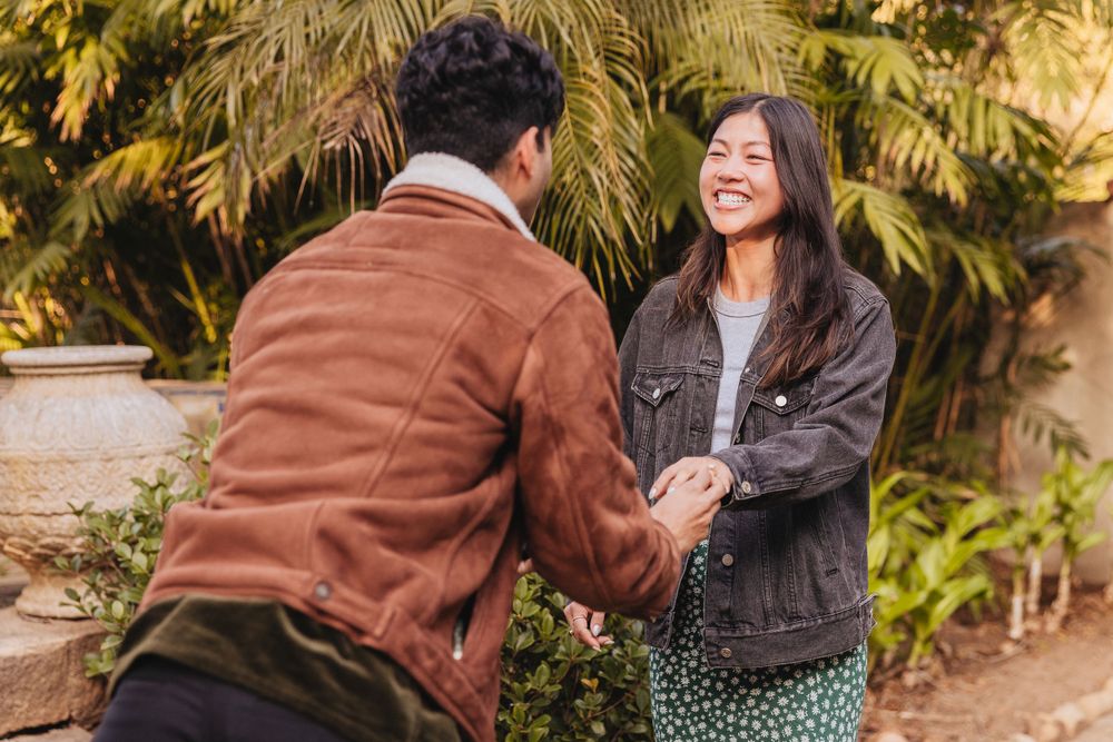 Two people holding hands and smiling while standing in a garden with palm trees in the background.