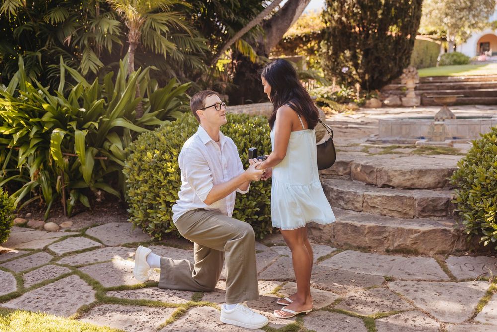 Man proposing on one knee to woman on garden steps surrounded by lush tropical greenery on a sunny day.
