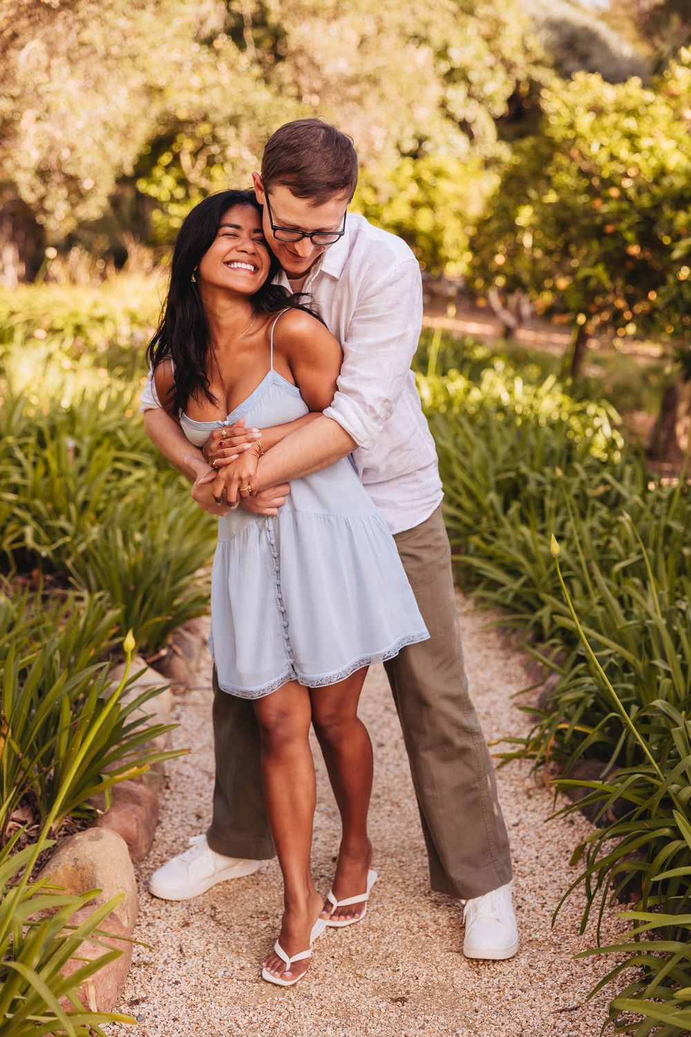 Happy interracial couple embracing on a garden path, woman in blue dress laughing joyfully in sunny outdoor setting.