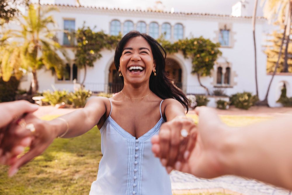 Joyful woman in blue sundress laughing while holding hands with someone in front of a white villa.
