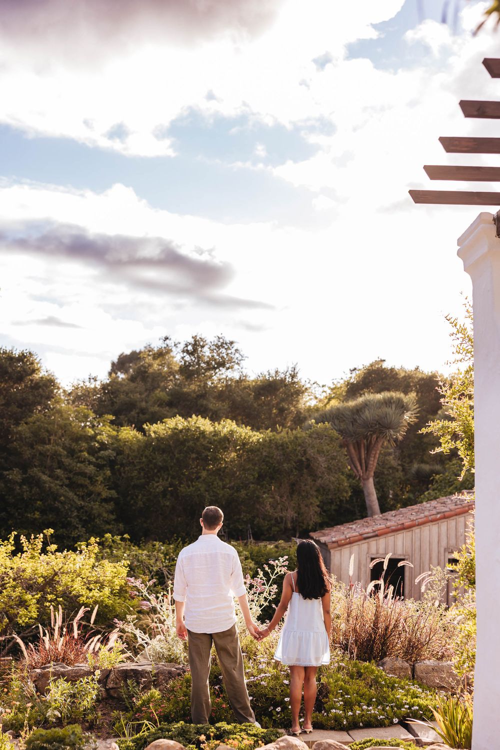 Couple holding hands from behind, overlooking a lush garden landscape with dramatic cloudy sky.