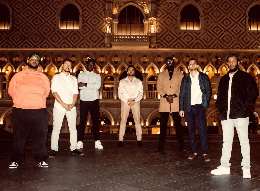 Seven men in stylish outfits pose inside an ornate golden-lit grand interior hall.