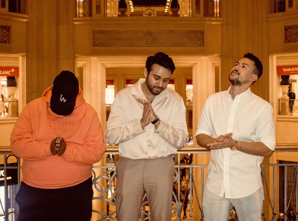 Three men posing indoors in an ornate golden lobby, one in orange, two in white shirts.