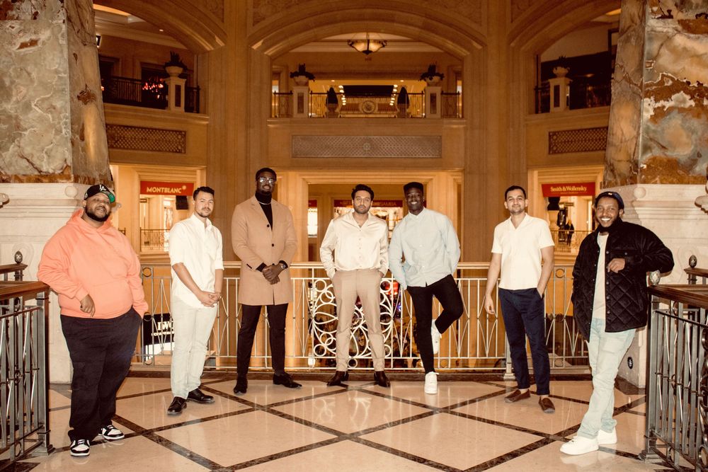 Group of seven men standing in a line inside a grand ornate hotel lobby.