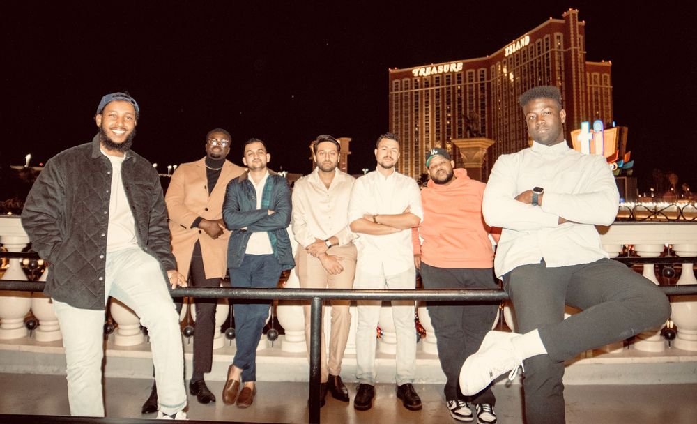 Group of eight friends posing on balcony with Mandalay Bay Las Vegas lit up at night.