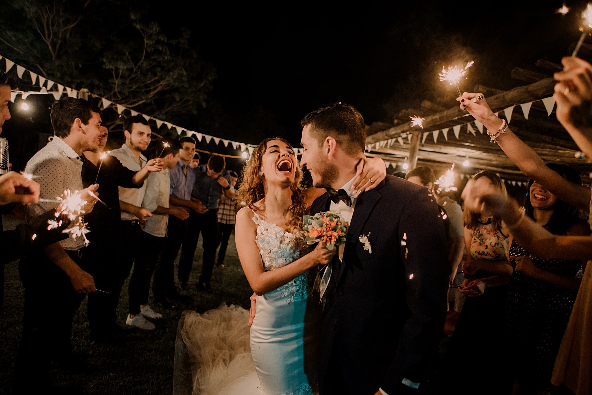 Bride and groom laughing joyfully surrounded by guests holding sparklers at an outdoor night reception. Fotógrafo en Formosa