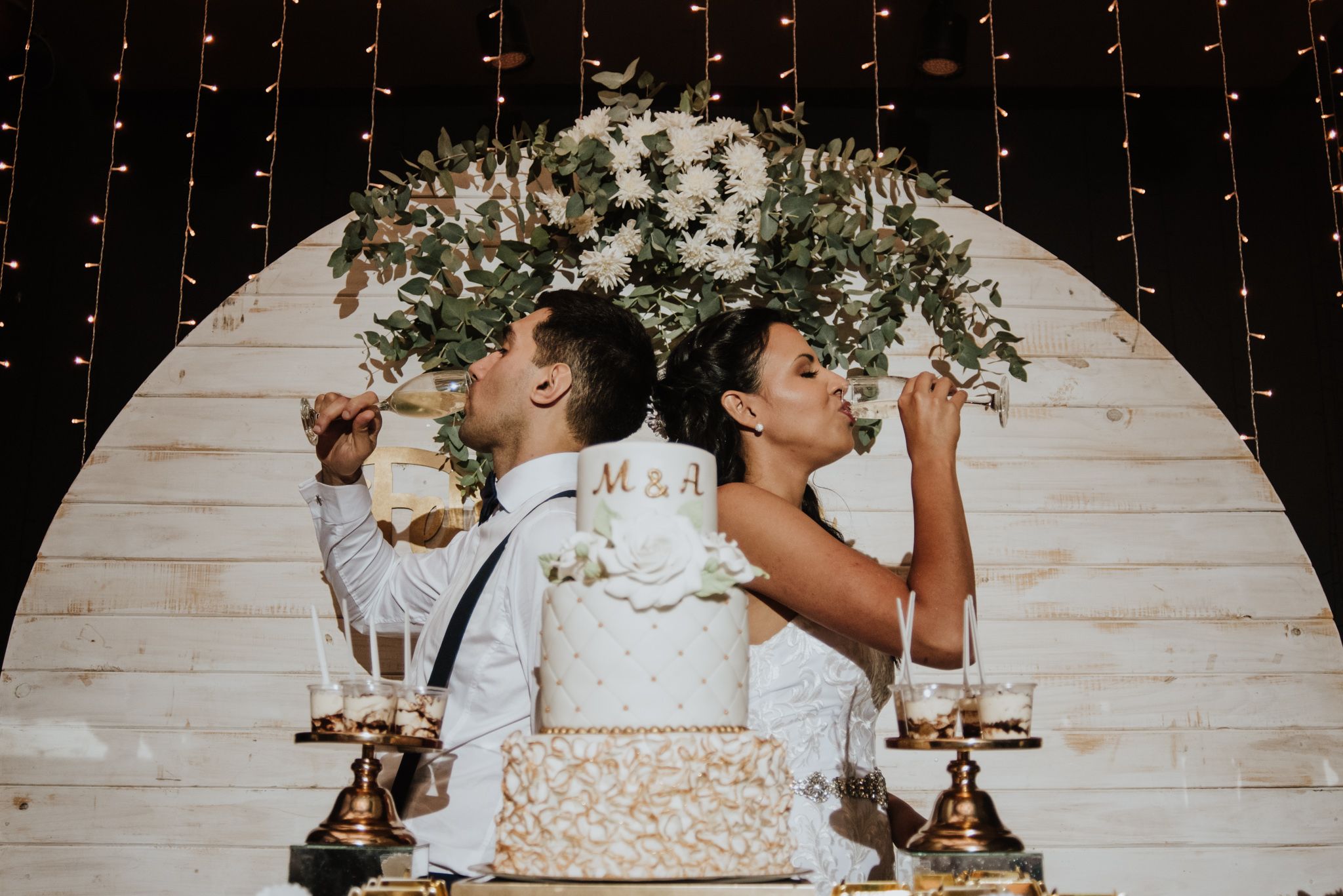 Bride and groom toasting back to back in front of an elegant wedding cake with floral backdrop. Fotógrafo en Formosa