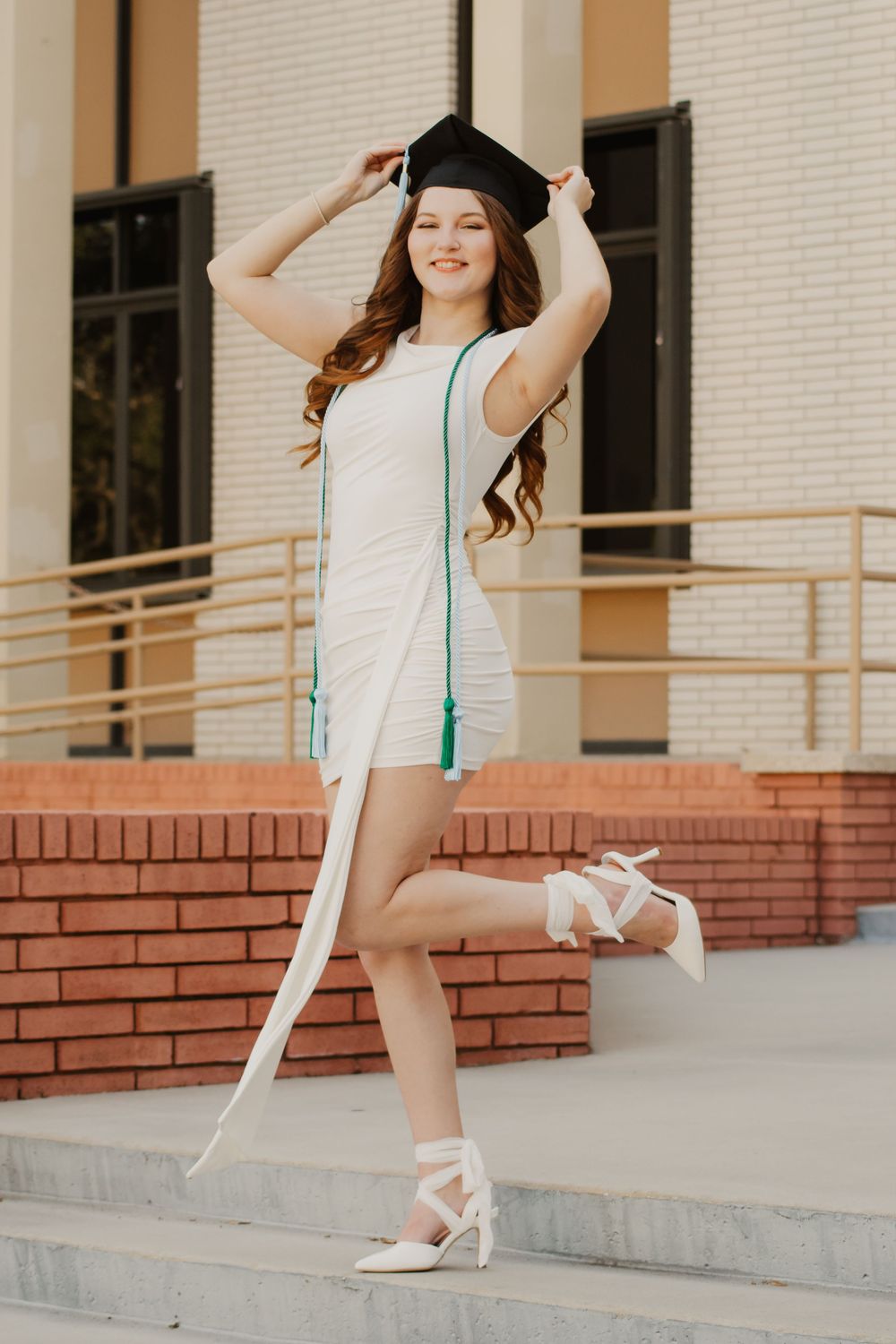 A graduate in white dress and graduation cap poses playfully on brick steps with her cap in hand.