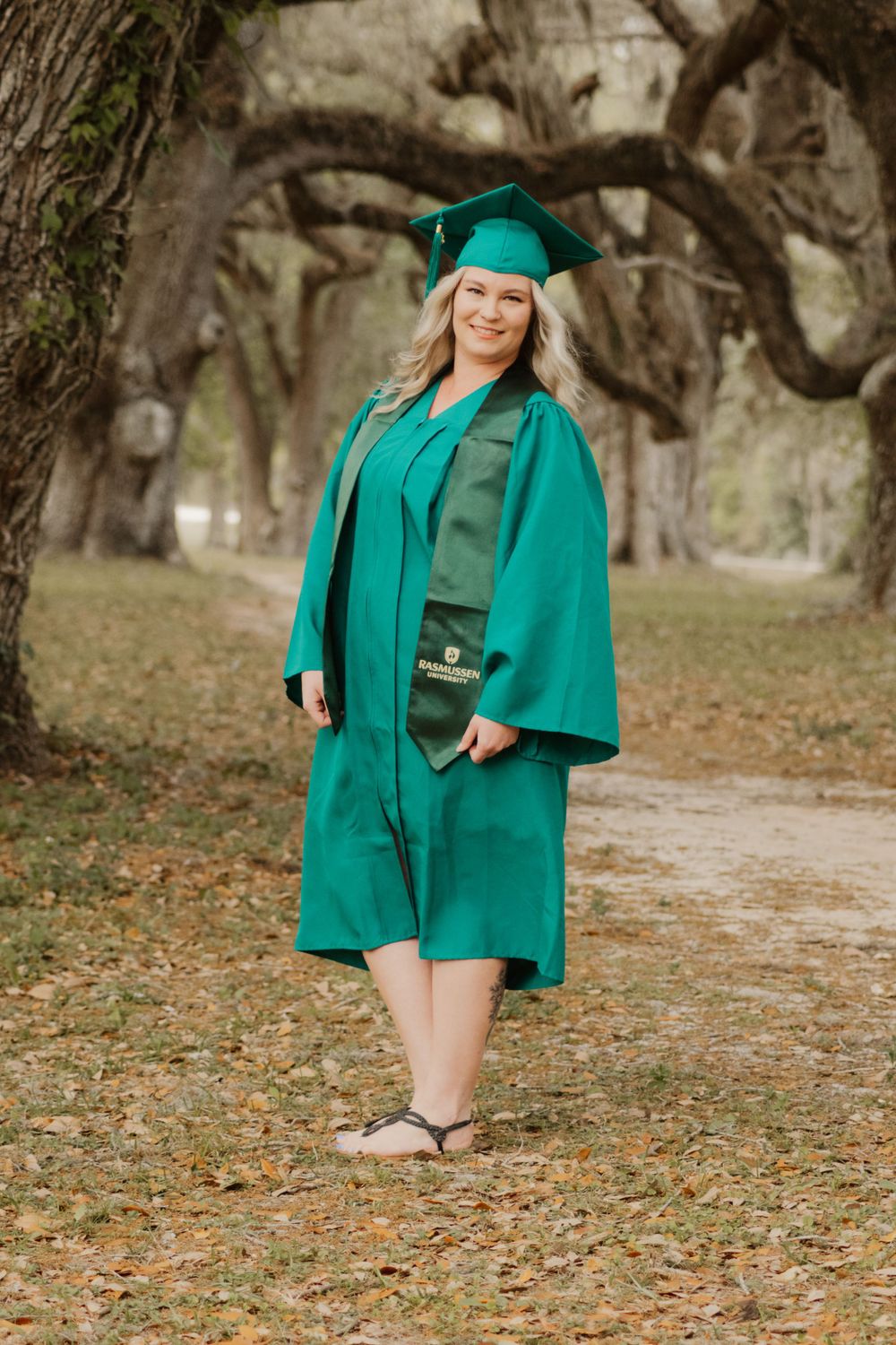 A graduate in emerald green cap and gown poses on a path covered with autumn leaves in a park setting.