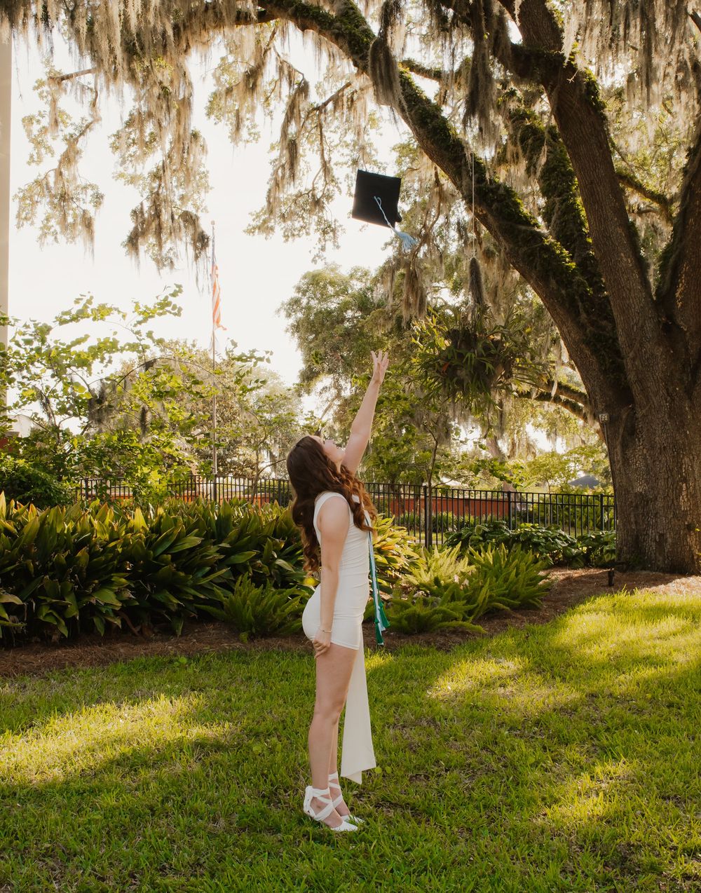 A graduate in white dress and graduation cap poses playfully on green grass while throwing her cap in the air.
