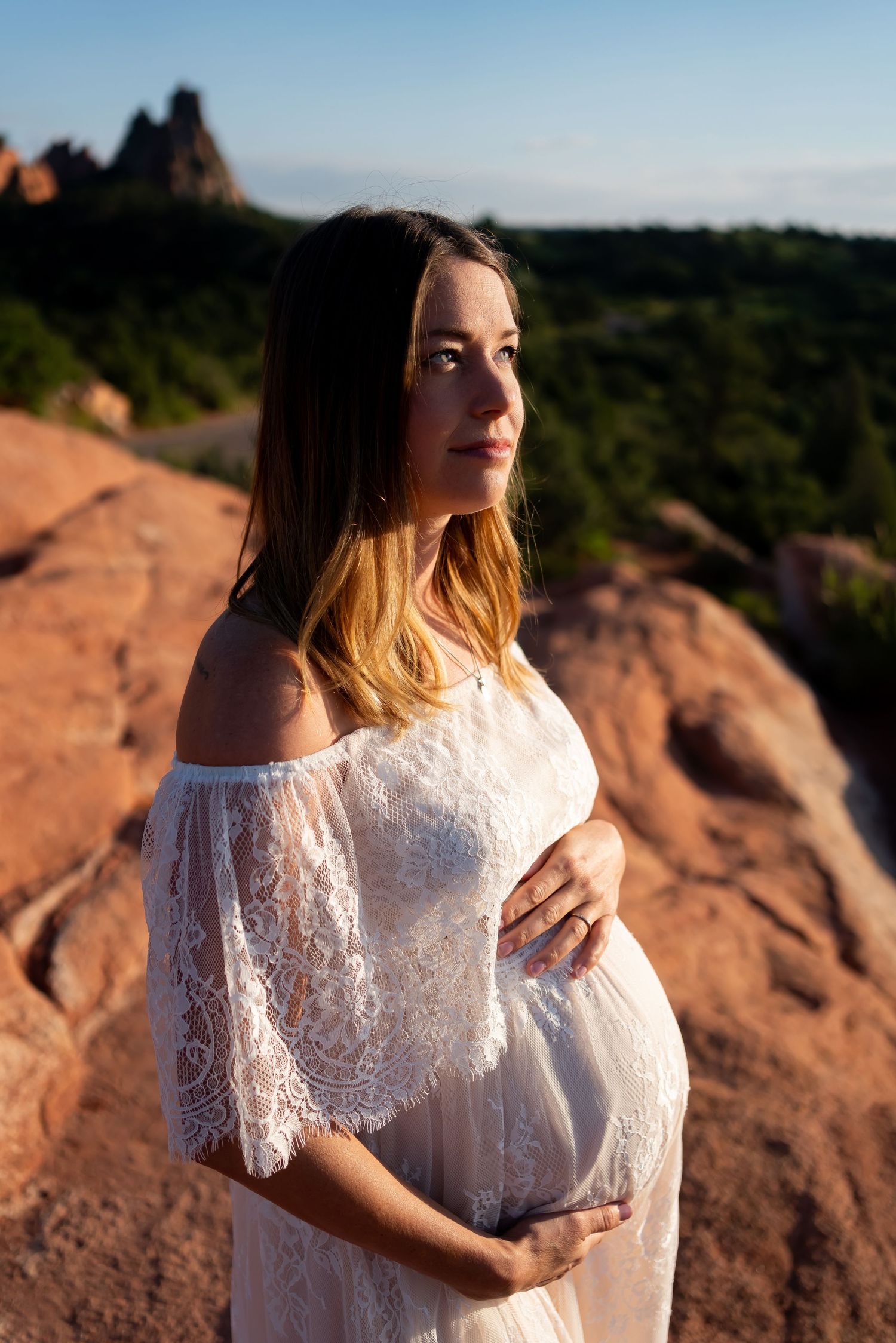 Expecting mother in white lace dress poses against red rock formations at sunset.