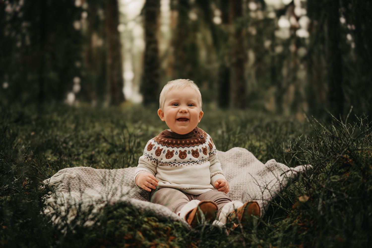 A happy baby in a Nordic sweater sits on a blanket surrounded by lush green forest undergrowth.