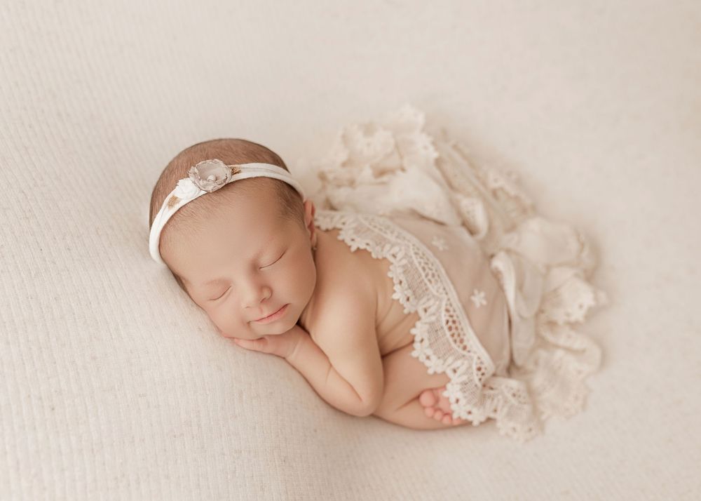 Sleeping newborn baby wrapped in delicate white lace fabric lies peacefully on cream colored blanket.