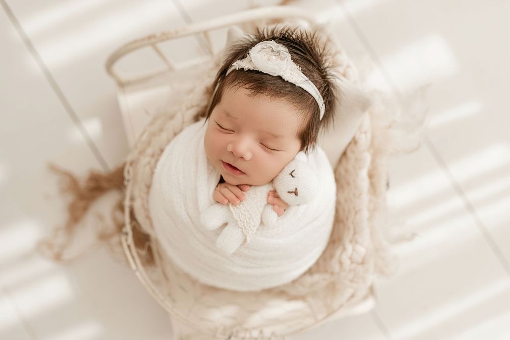 Newborn baby in white dress sleeps peacefully on soft white blanket during professional photo session.