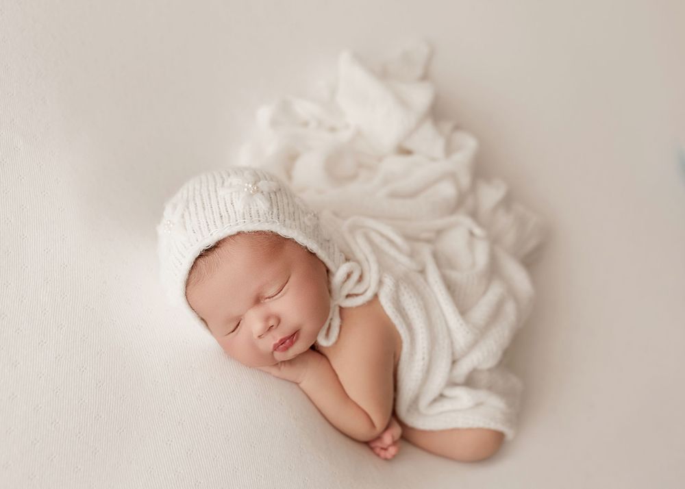 A sleeping newborn baby wrapped in soft white fabric lays peacefully against a light beige background.