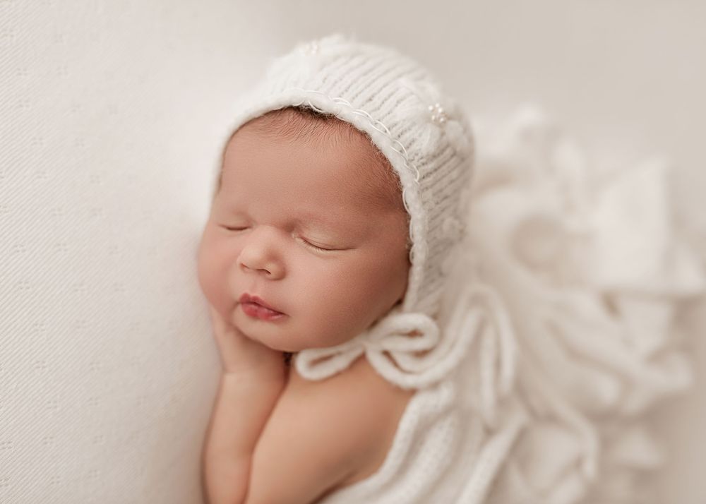 Peaceful sleeping newborn wearing soft white knit bonnet rests on cream colored blanket in gentle lighting.
