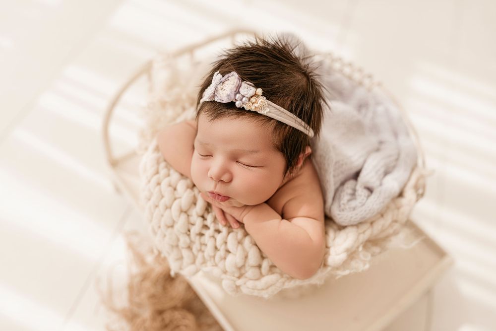 A sleeping newborn baby wrapped in delicate lace fabric with a dainty floral headband rests on a cream background.