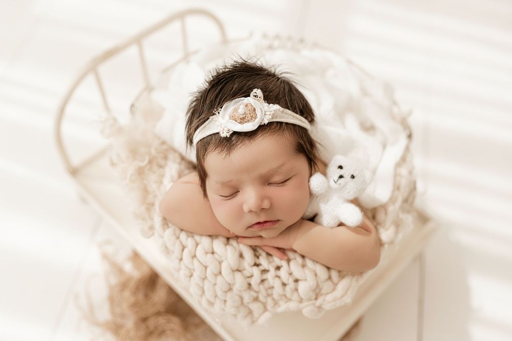 A sleeping newborn baby wearing a delicate white headband lies peacefully on soft cream-colored fabric.