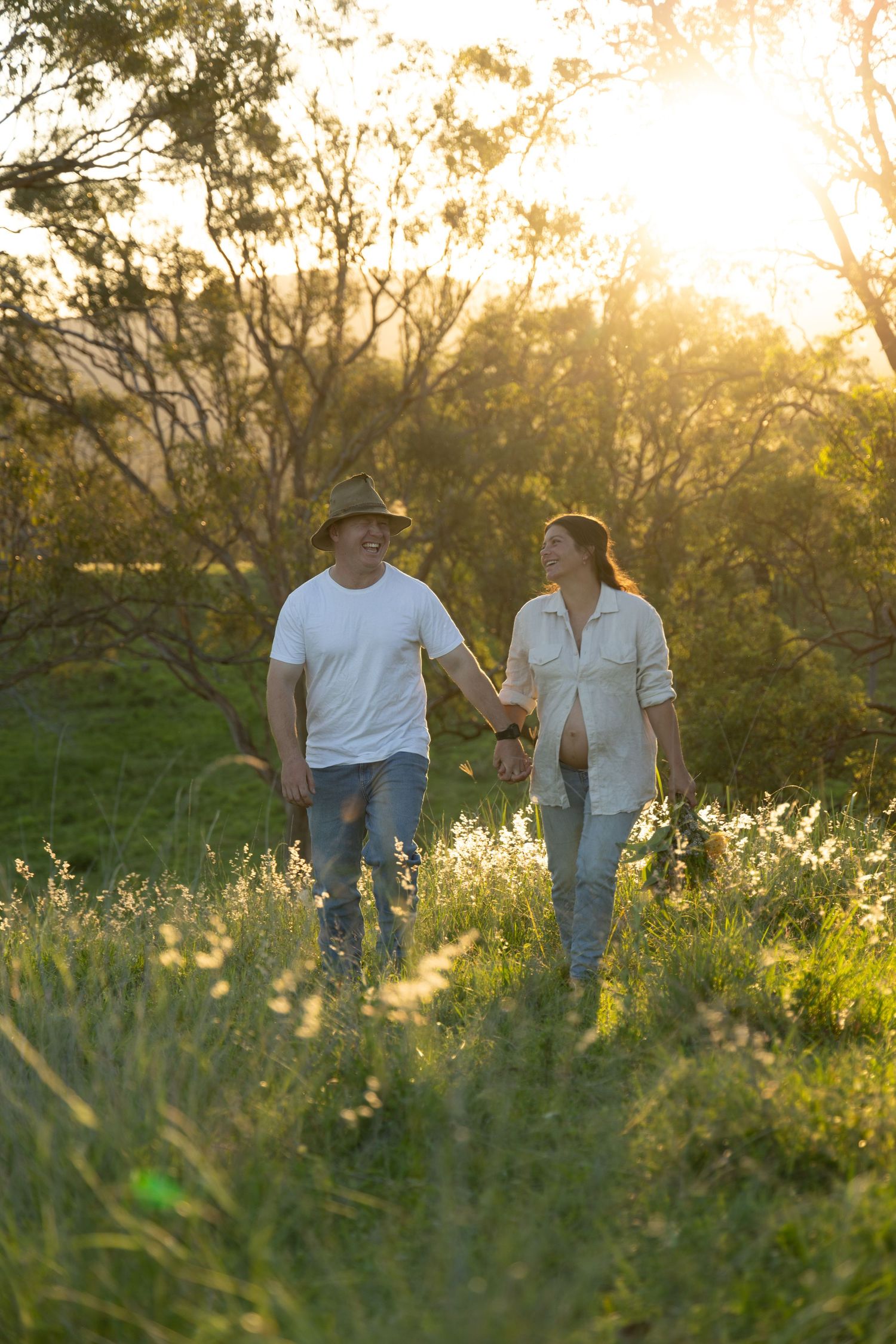 A couple who are expecting walks together through a sun-drenched meadow during golden hour as warm light filters through trees.