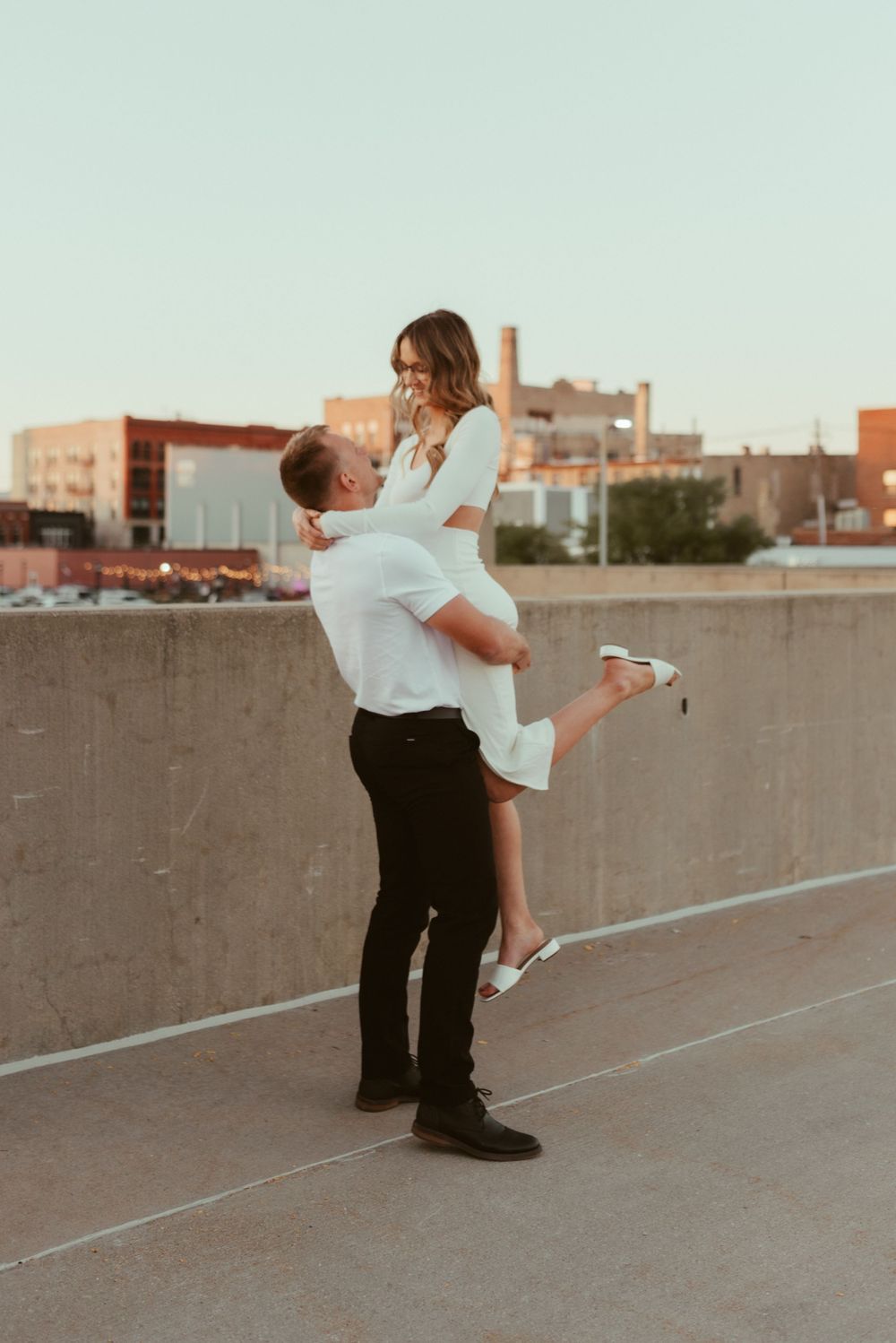 Man lifting woman in white dress during romantic rooftop photoshoot.