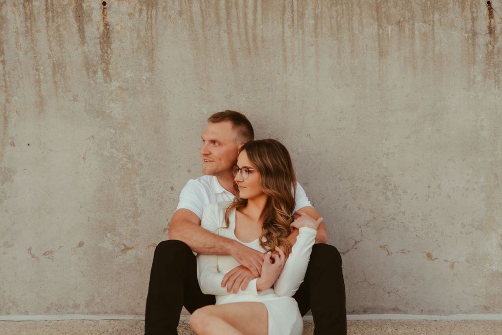 A couple in formal attire sits close together against a concrete wall background.