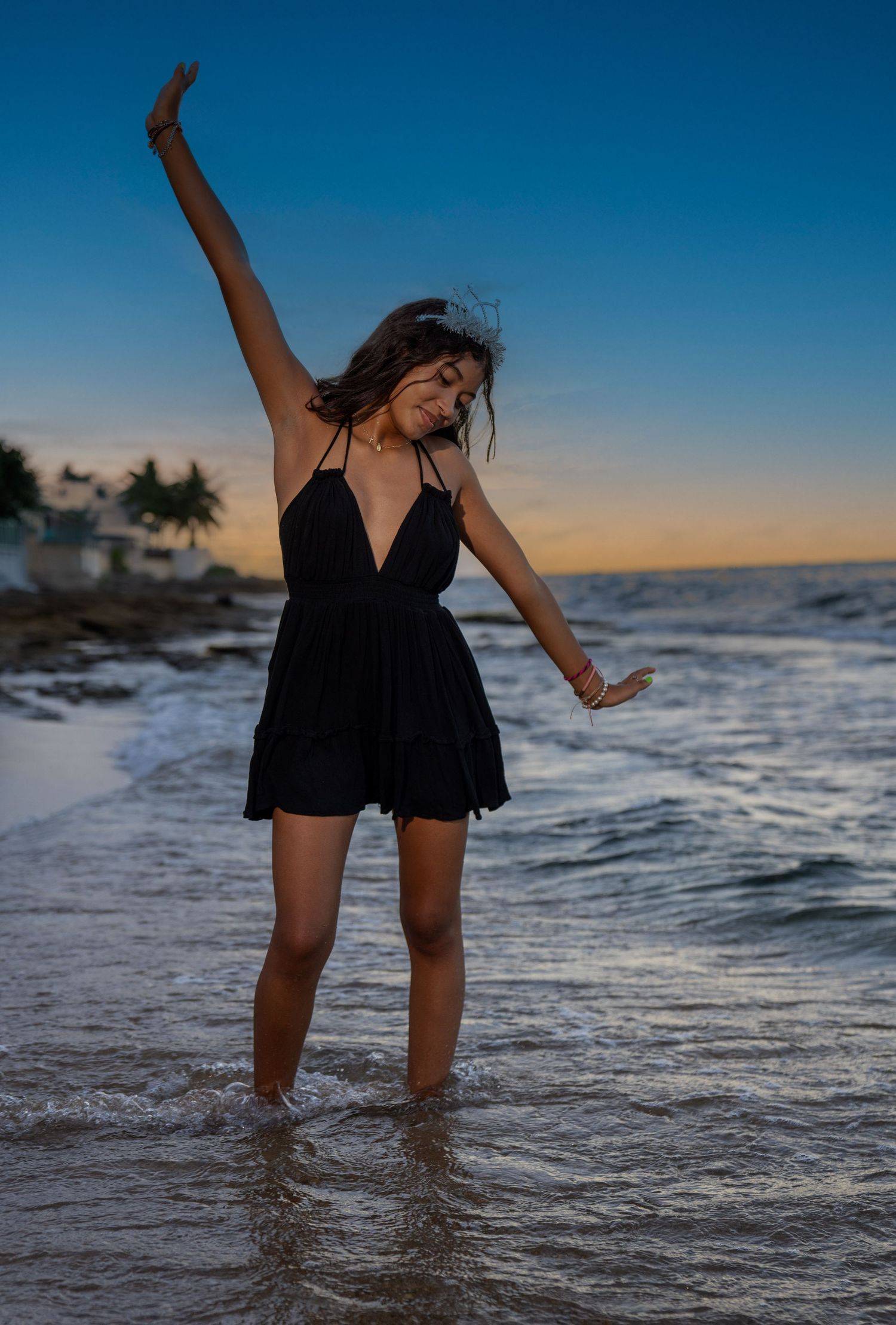 Woman in black dress dancing at sunset on beach with waves crashing.